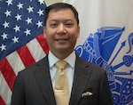 Smiling man in dark gray suit stands in front of flags