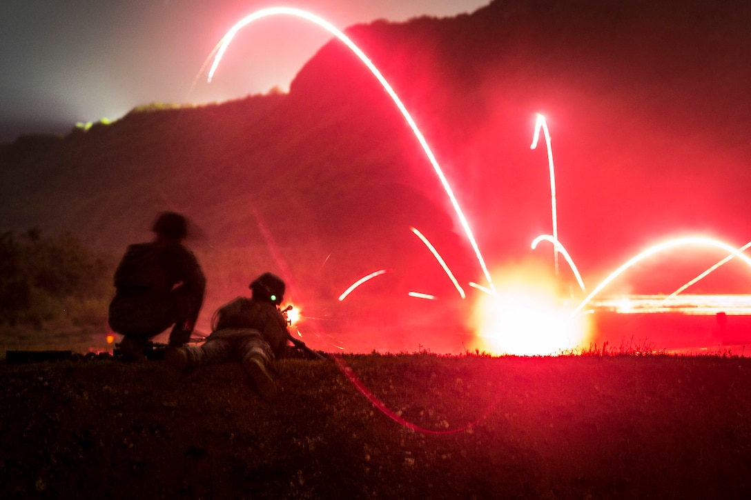 Two Marines fire their weapons causing tracers in a red sky.
