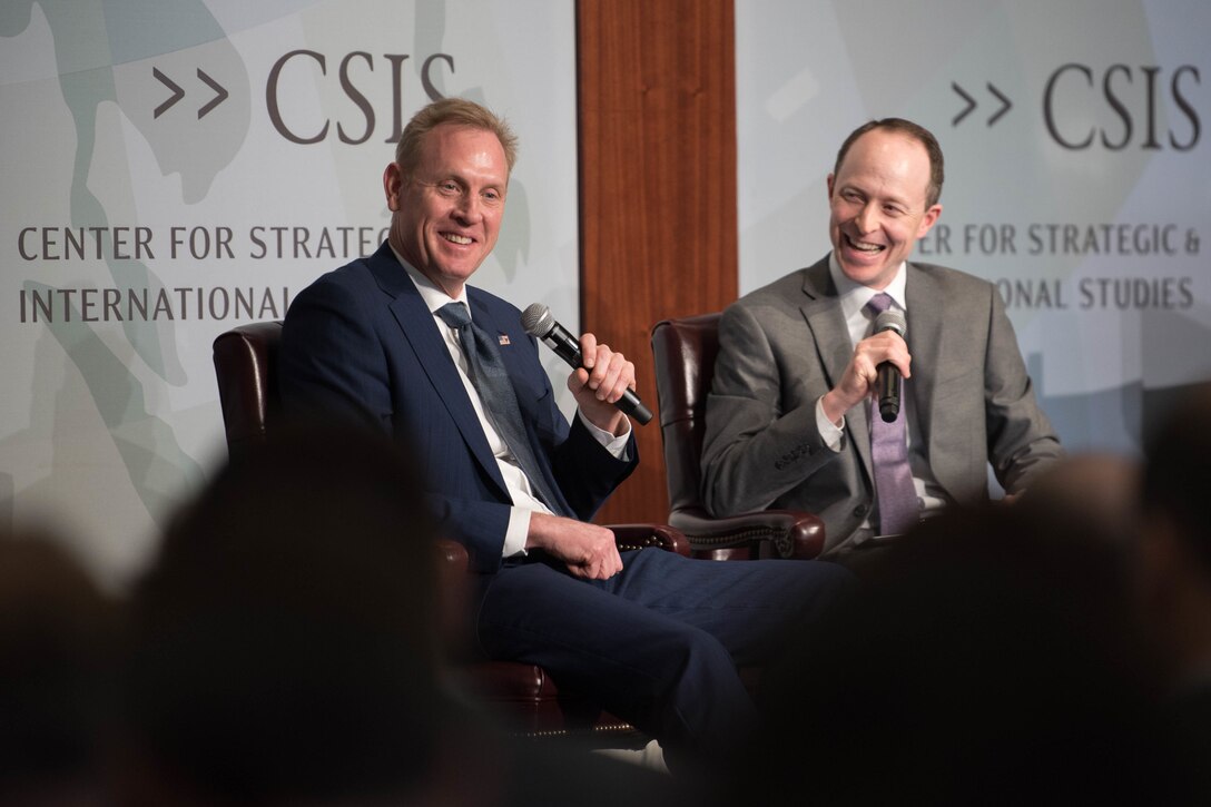 Acting Defense Secretary Patrick M. Shanahan sits on a stage holding a microphone next to another man sitting on stage holding a microphone.