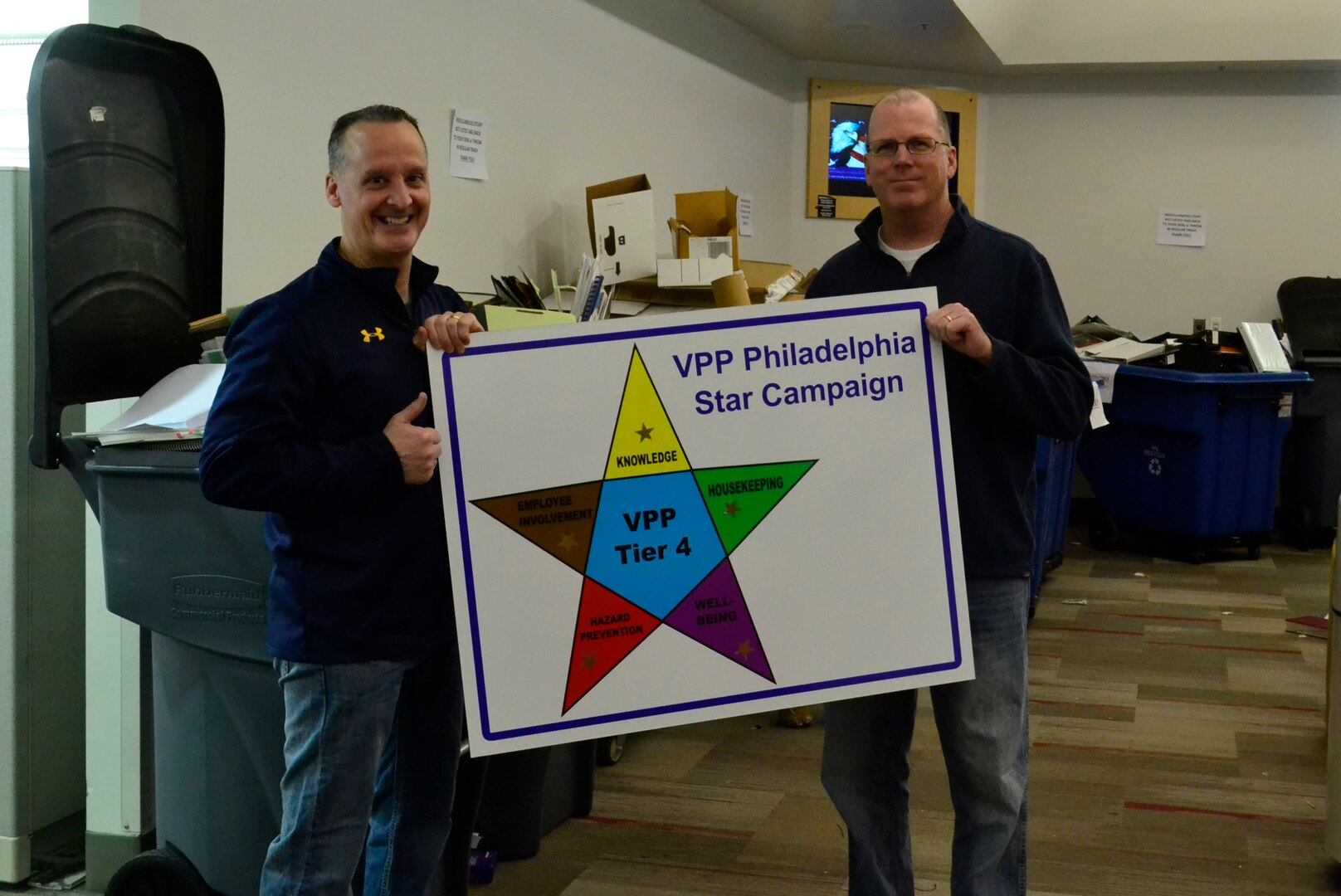 Navy Capt. Gerald Raia, (left), Construction and Equipment supply chain director, and Tom Grace, (right) Construction and Equipment deputy director hold the VPP Star campaign placard during the spring clean-up event in Building 3 March 20, 2019 in Philadelphia. The leaders implemented a supply chain wide housekeeping event to demonstrate the directorate’s commitment to the VPP program, an OSHA led initiative that promotes good housekeeping in the office environment as a major part of its mishap, pest management and fire prevention efforts. (Photo by Alexandria Brimage-Gray).