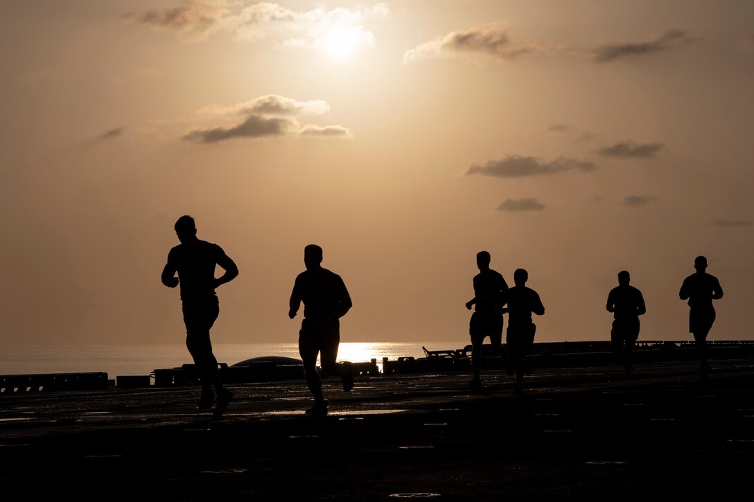 Six men are silhouetted against the sky as they run along the flight deck of a military ship.