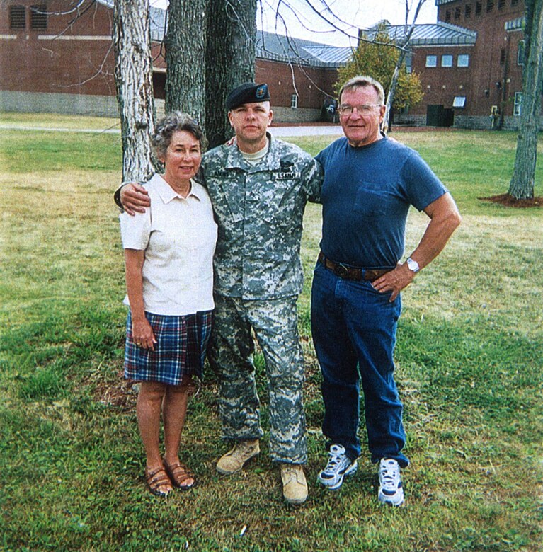 A soldier stands with arms around his parents in front of a tree.