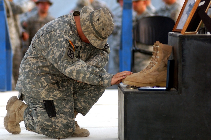 A soldier kneels with head down in front of a memorial that includes a boot and photo frames.