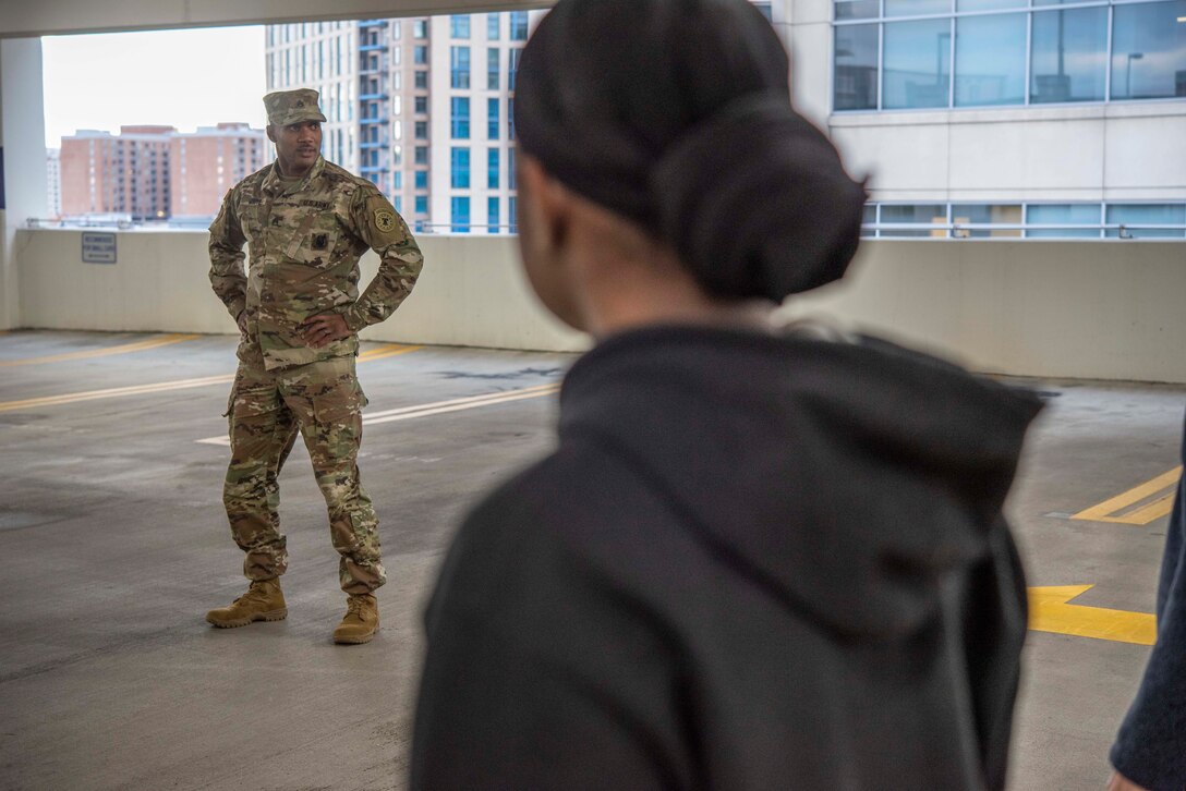 A soldier in camouflage stands with his arms akimbo in a parking garage.