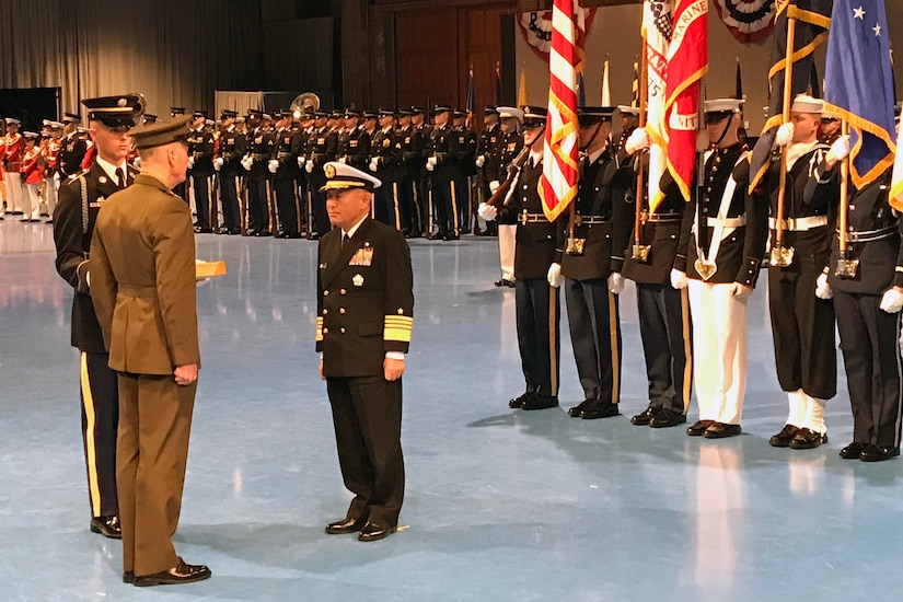 Two military men face each other as a military color guard stands behind them.