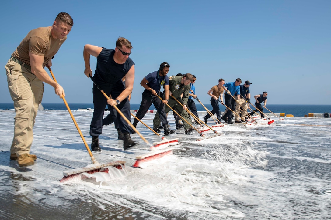 A line of men use brooms to clean the flight deck of a military ship.