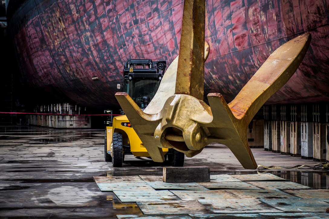 A ship's anchor sits in a dry dock with a construction vehicle parked behind it.