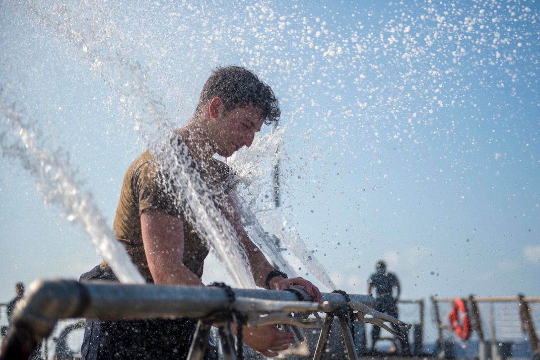 A sailor holds onto a pipe as water bursts onto his face.