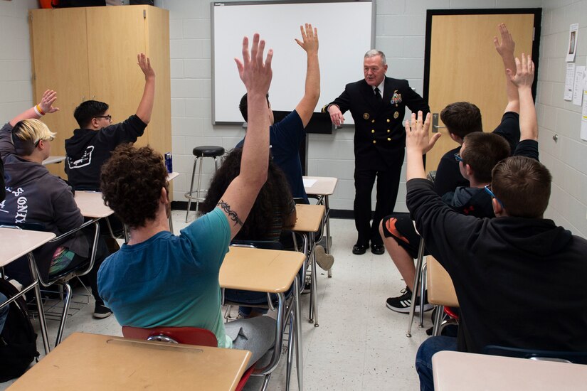 A sailor stands at the front of a classroom full of students with their hands up.