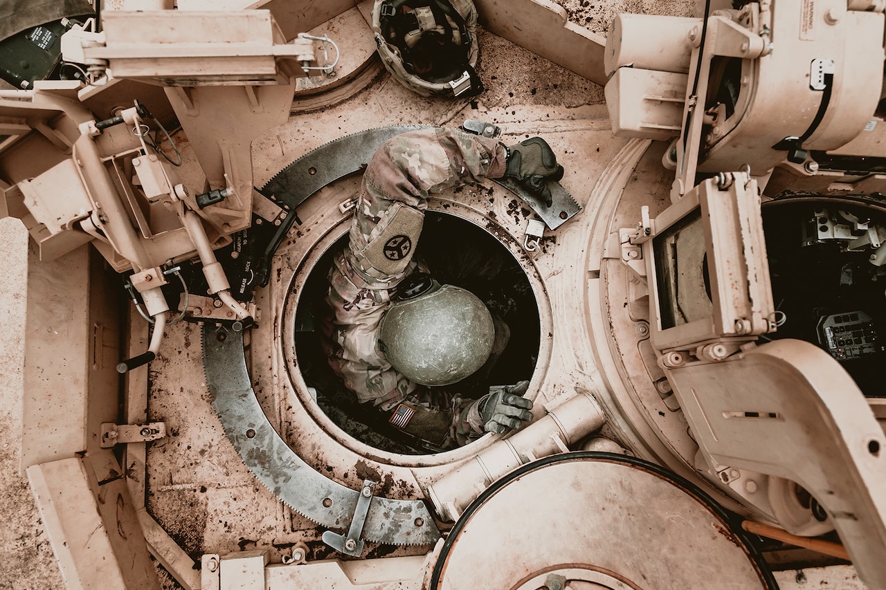 A soldier, shown from overhead, emerges from the top opening of a tank.