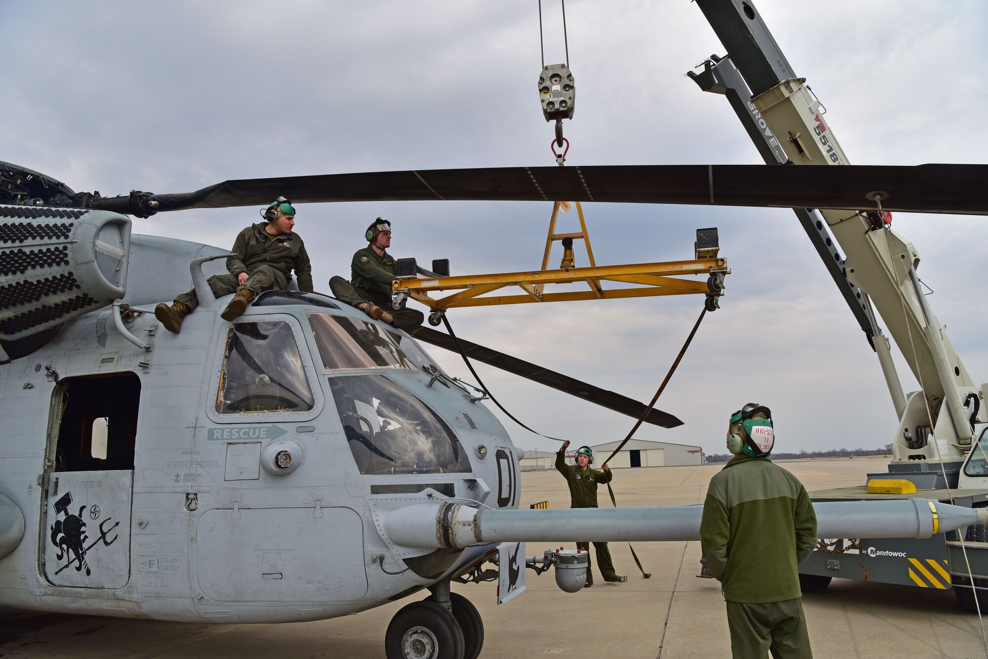 Men wearing a flight suit prepare to put a rotor blade from a CH-53E Super Stallion onto a crane.