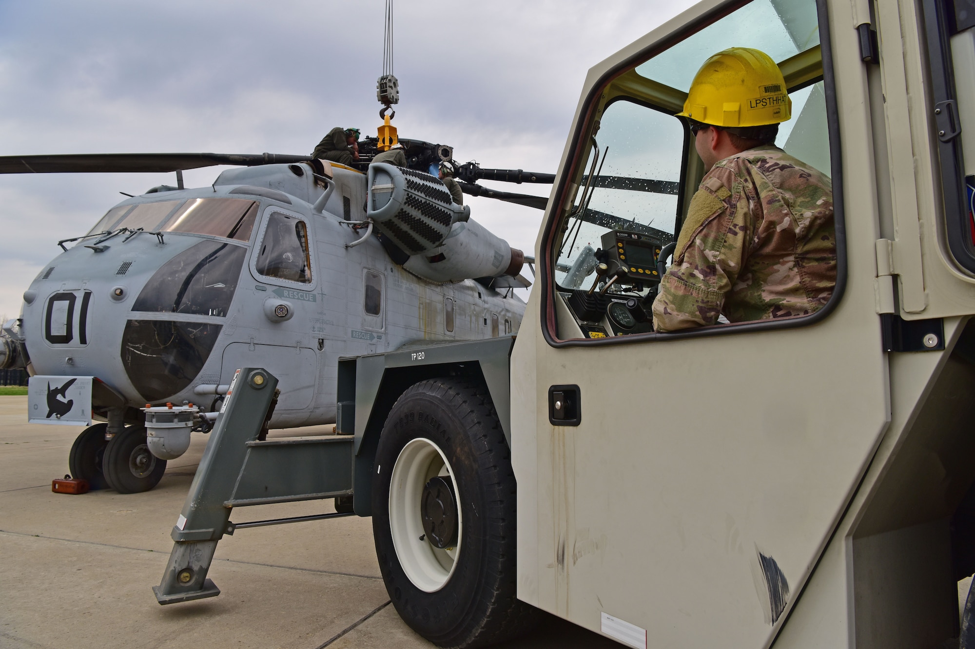 A man wearing the operational camouflage pattern uniform sits in a crane in front of a CH-53E Super Stallion.