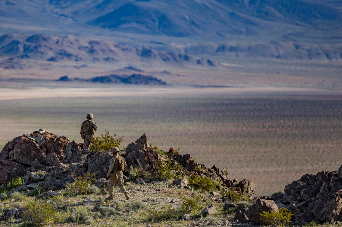 Two Marines walk through dessert with a mountain in the background.