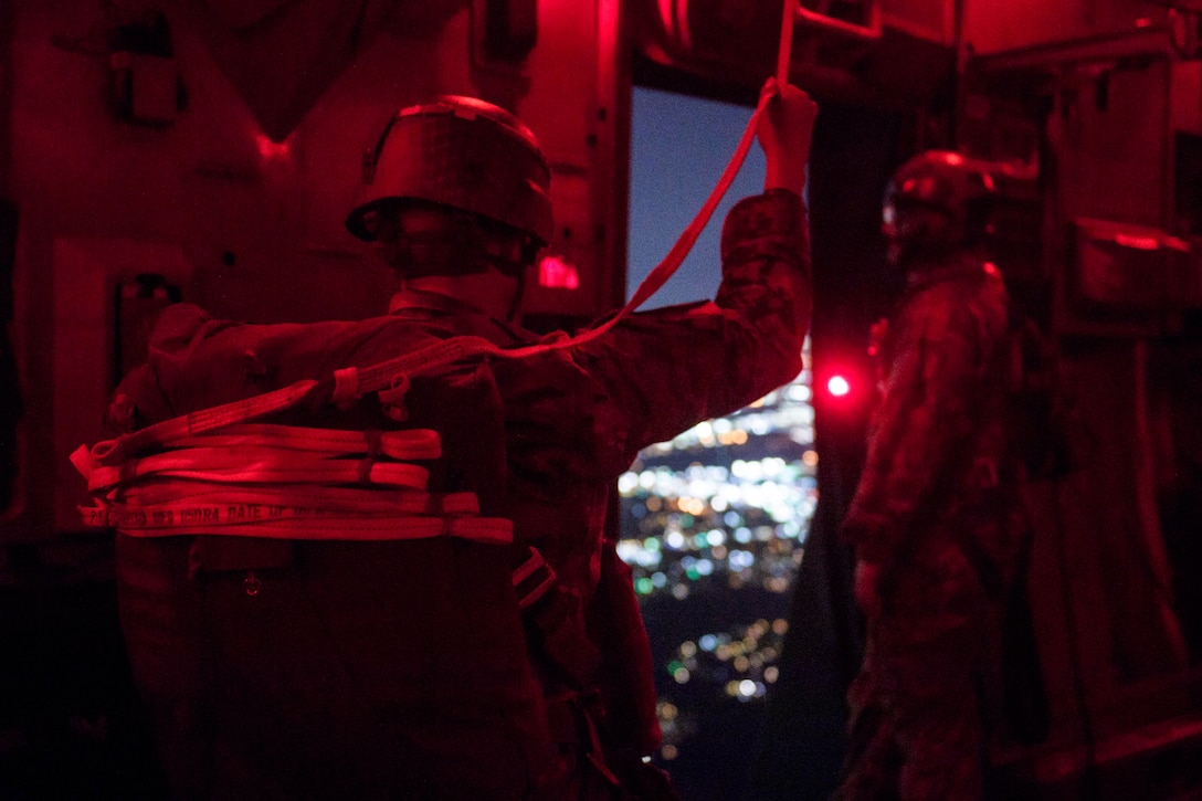 A soldier stands in front of the door of an aircraft looking out into the sky surrounded by a haze of red light.