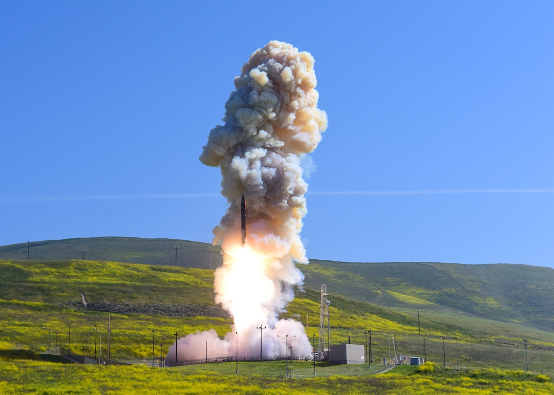 A missile launches into blue sky from a launch pad surrounded by rolling green fields.