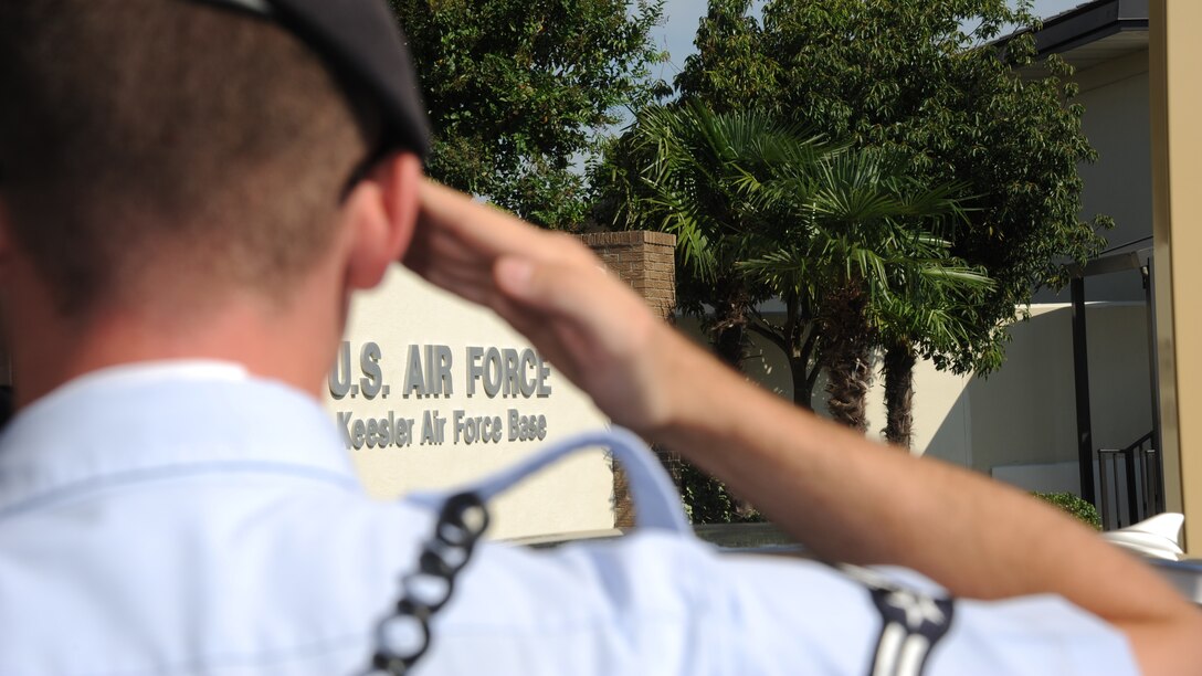 A view from behind and over the shoulder of a saluting airman with the Keesler Air Force Base sign in the background