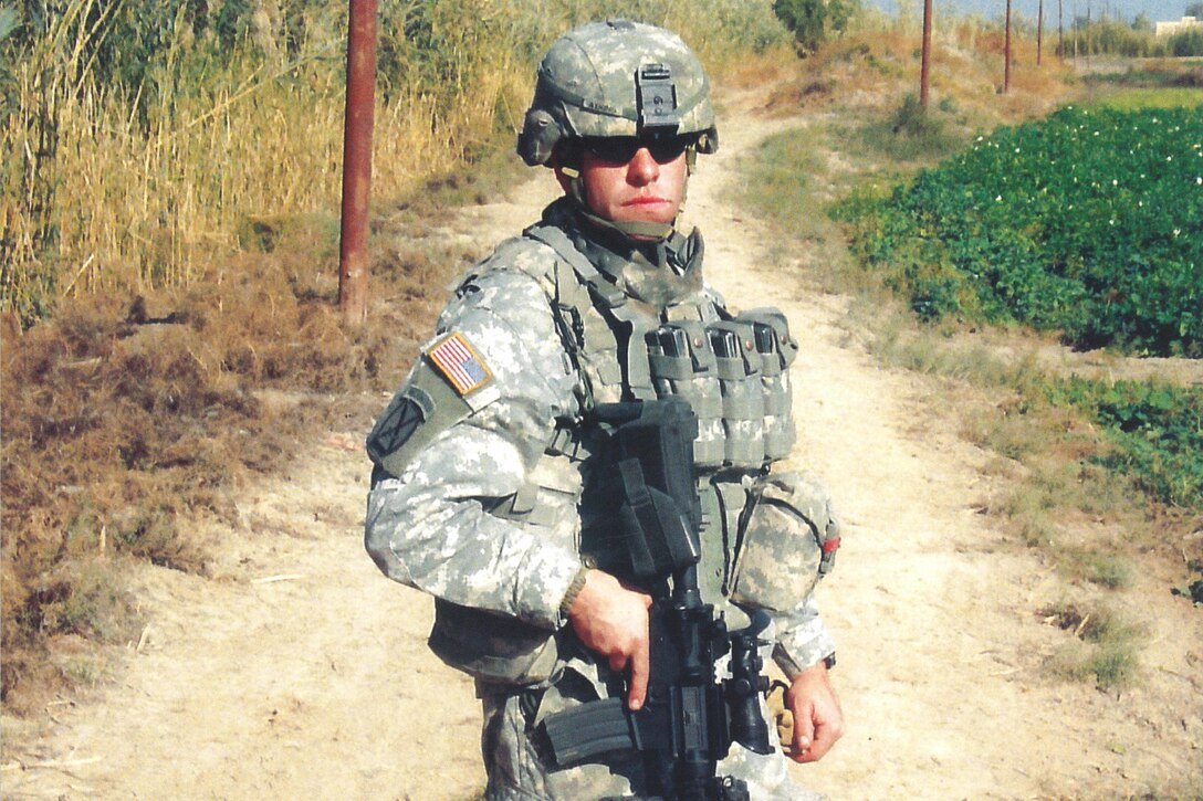 A soldier wearing sunglasses stands on a dirt road holding a weapon.