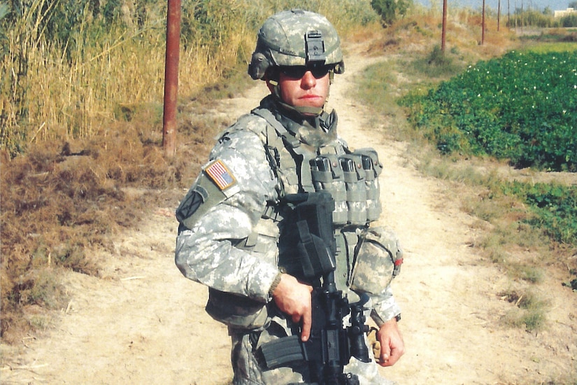 A soldier wearing sunglasses stands on a dirt road holding a weapon.