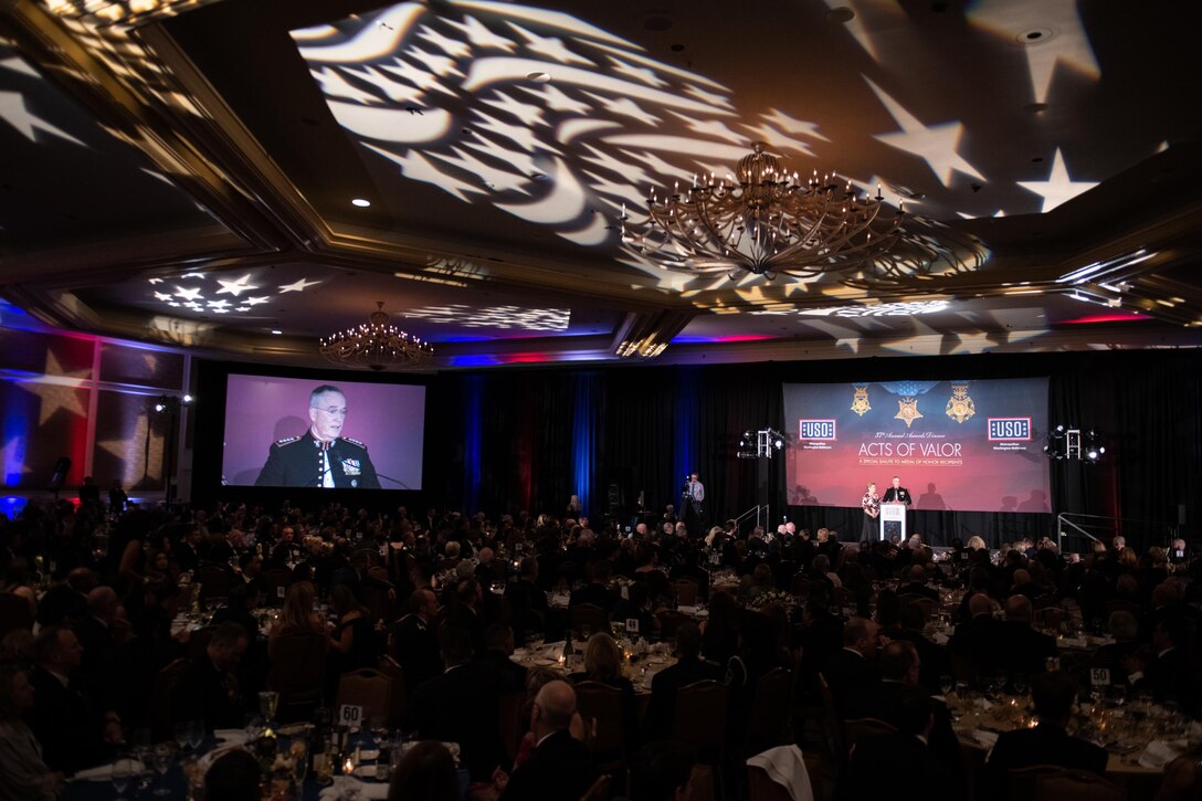 Large screen in banquet hall shows Joint Chiefs chairman speaking from a lectern