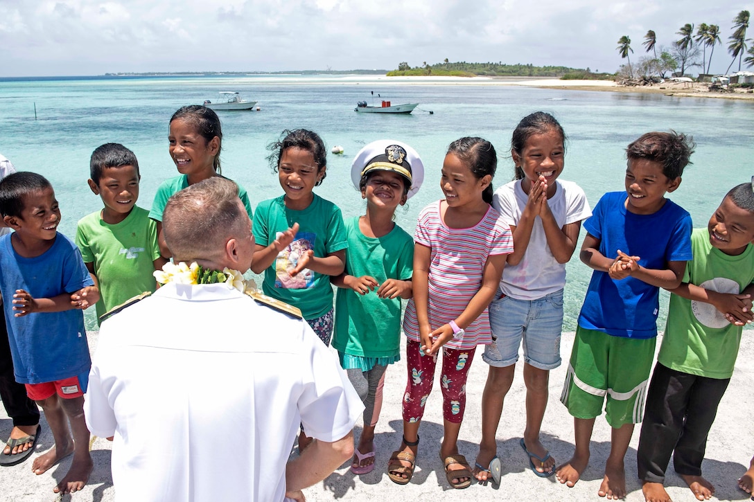 A Navy commander speaks to a group  of children on a beach.