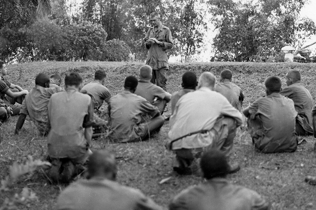 Several men sit in a field watching a man standing up reading.