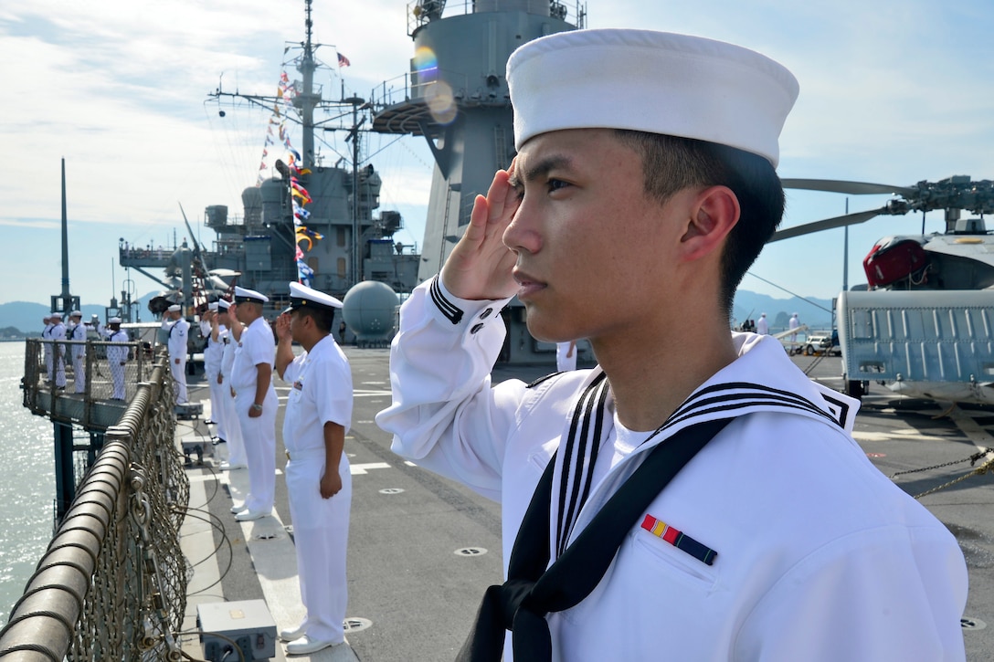 Sailors salute while standing along the rail of a ship.