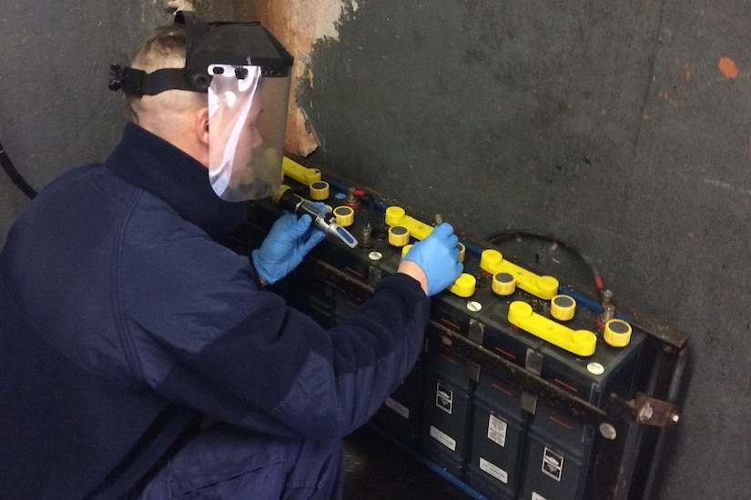 A Coast Guardsman in a blue jumpsuit works on a rack of large batteries.