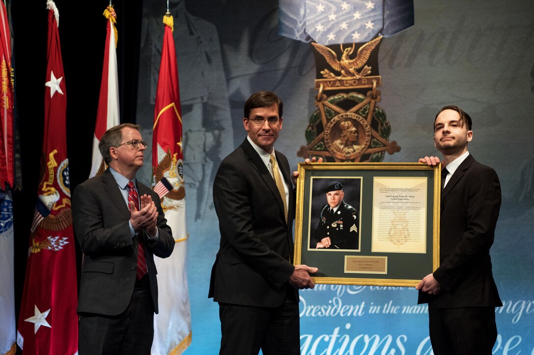 David L. Norquist applauds on a stage as two men beside him hold up a framed photo and certificate.