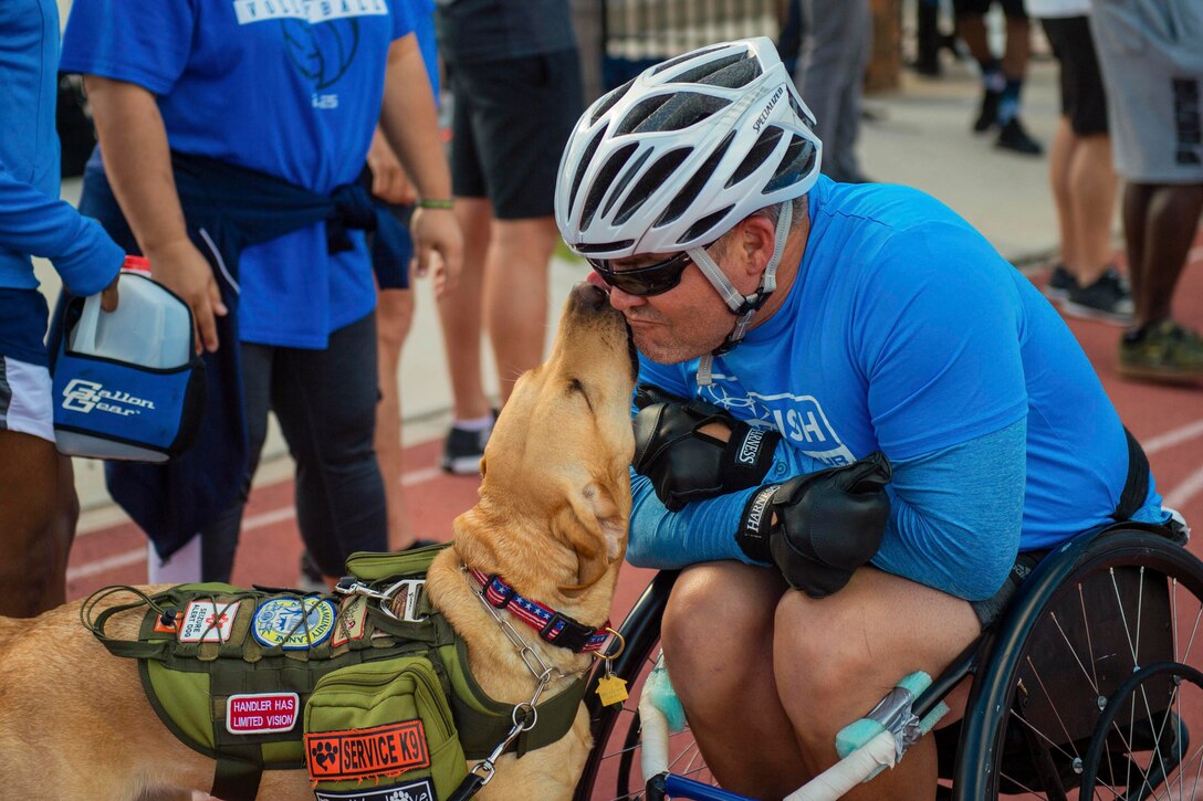 A sailor sits in a wheelchair while a dog licks his face.