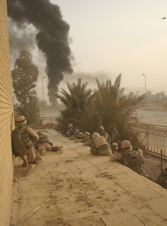 Soldiers huddle near a building with a plume of black smoke in the distance.