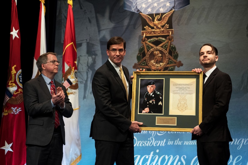 David L. Norquist applauds on a stage as two men beside him hold up a framed photo and certificate.