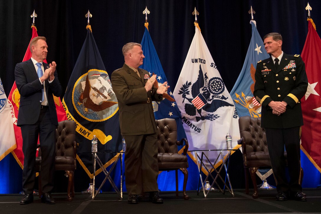 Three men stand on a stage with two of them applauding.