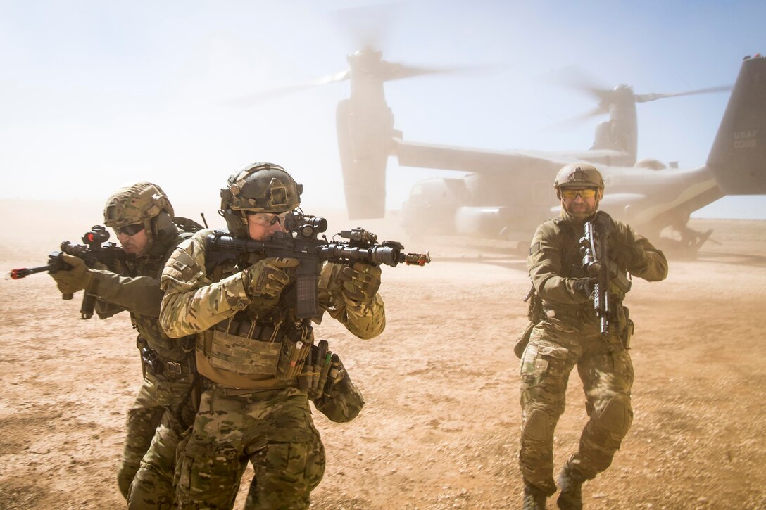 Three service members point rifles in various directions as they move through dust kicked up by an aircraft.