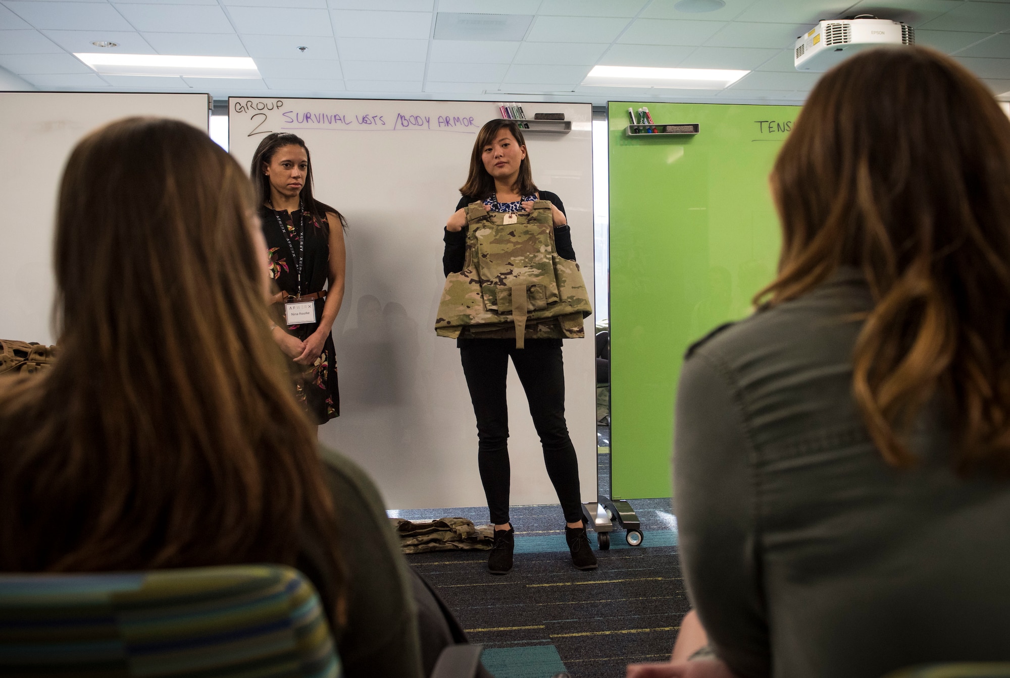 A woman holds up body armor and displays it to other women.