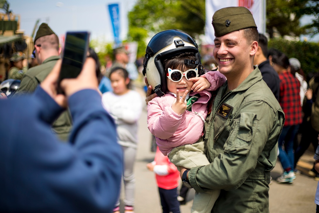 A Marine holds a child  who is wearing a helmet and sunglasses as a person takes their picture with a cellphone.