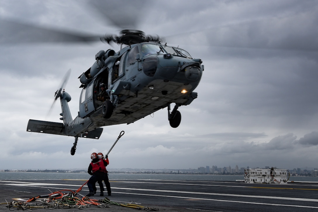 Two sailors attach slings to a helicopter while it flies above a military ship.