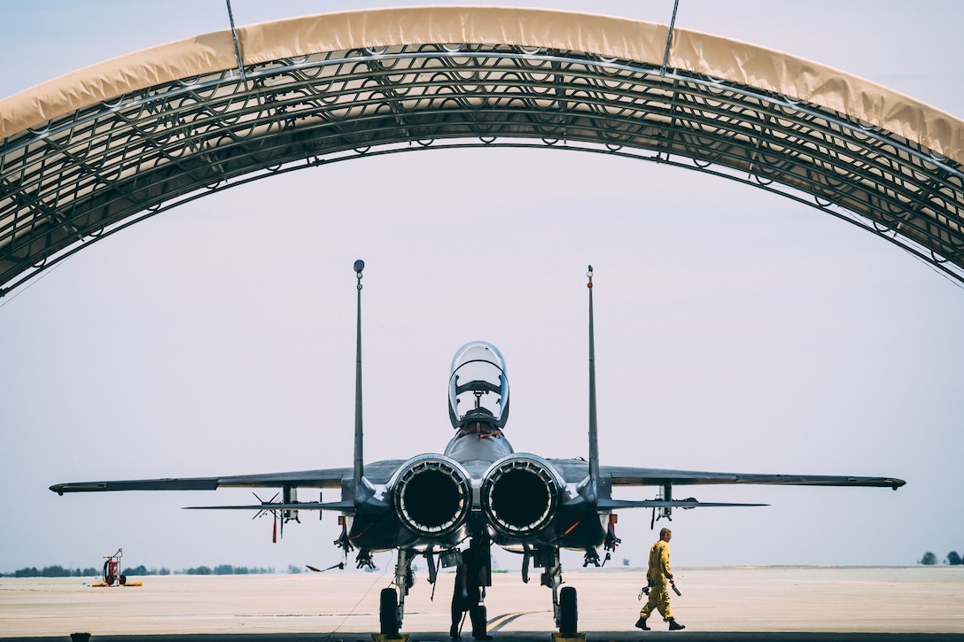 A fighter jet is shown from behind under a canopy as one airman checks the underside and another in a yellow jumpsuit walks off.