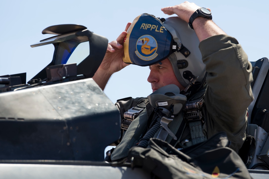 A man wearing a flightsuit and helmet sits in the cockpit of a fighter jet and pulls a colorful visor over his eyes.