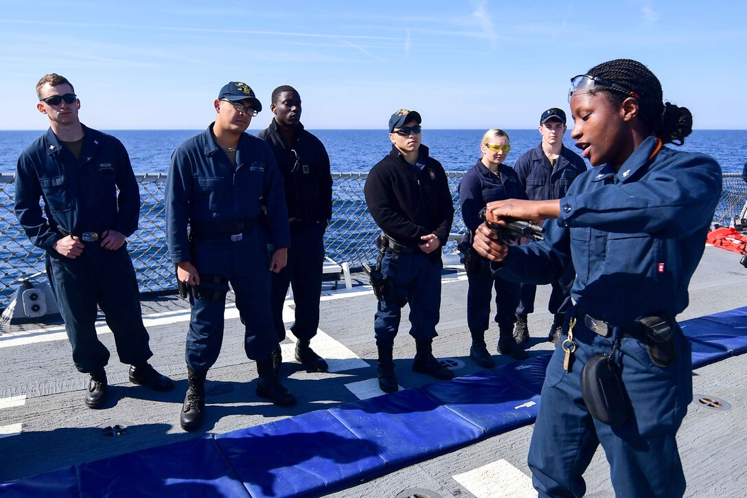 A sailor holds a gun as she talks to other sailors on a ship.