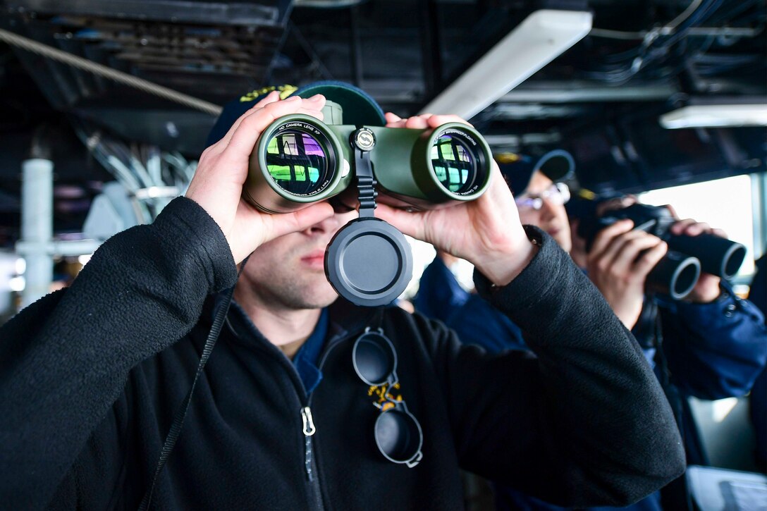 A sailor looks through binoculars while aboard a ship.