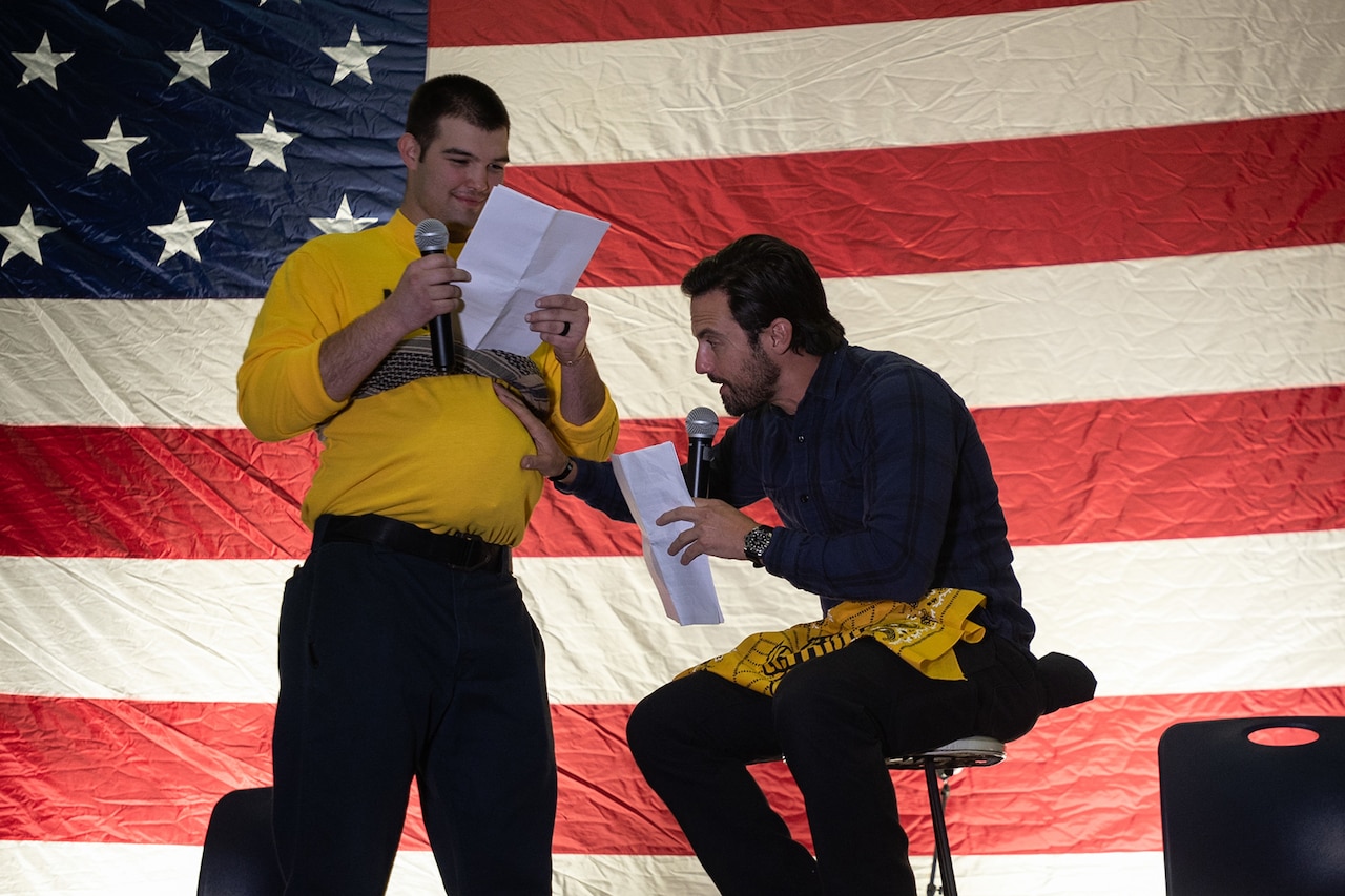 A man sits on a stool holding a microphone and the fake pregnant stomach of a man standing and reading from a script beside him. A U.S. flag is the backdrop.