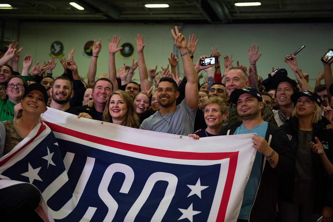 A group of people holding a USO banner raise their hands and give peace signs to a camera.