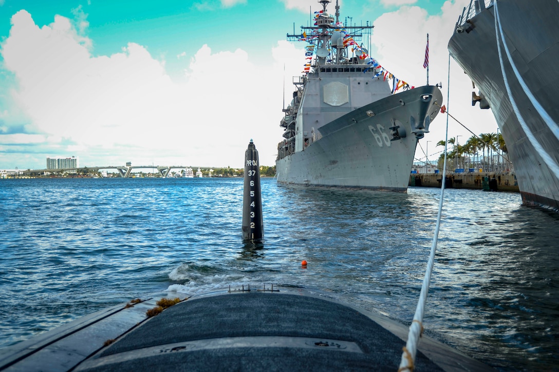 A submarine slightly above water attached by a rope to a large ship.