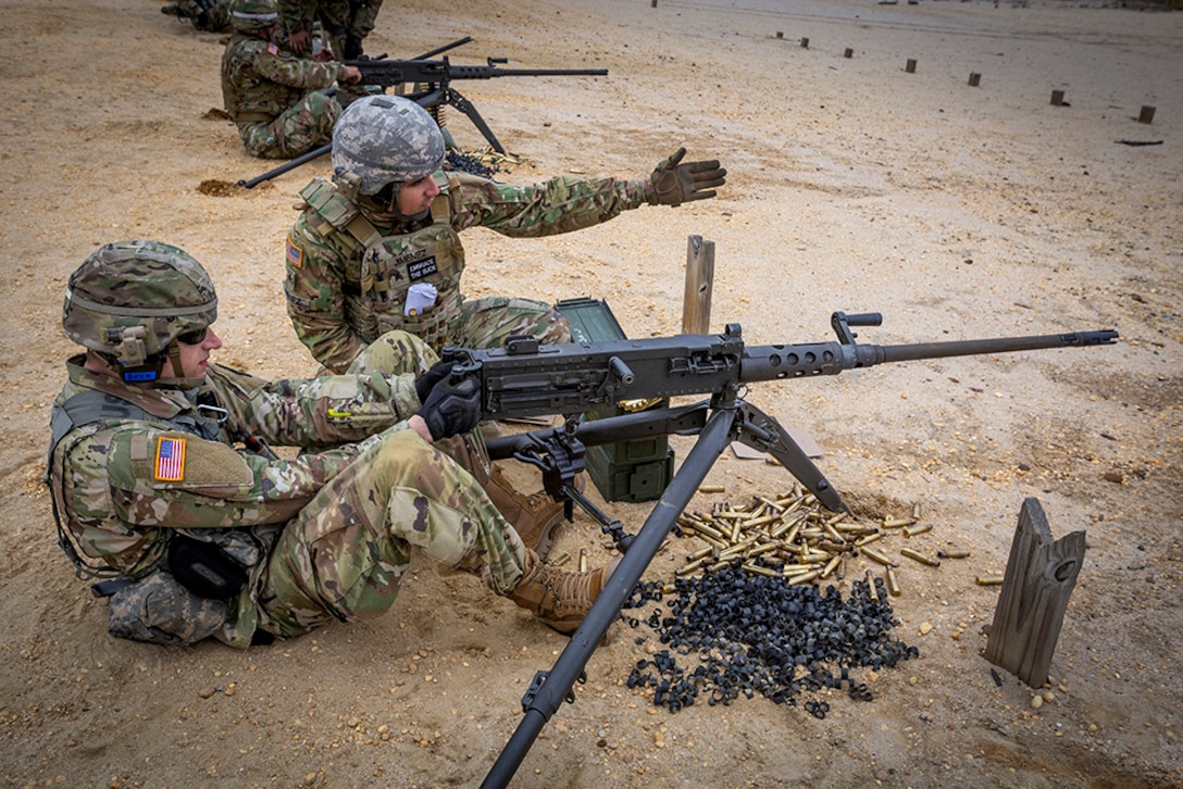 A soldier sits on the ground in front of a machine gun while another soldier sits beside him.