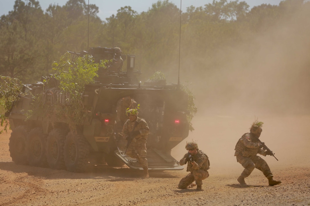 Soldiers stand and kneel around a military vehicle as dust swirls around them.