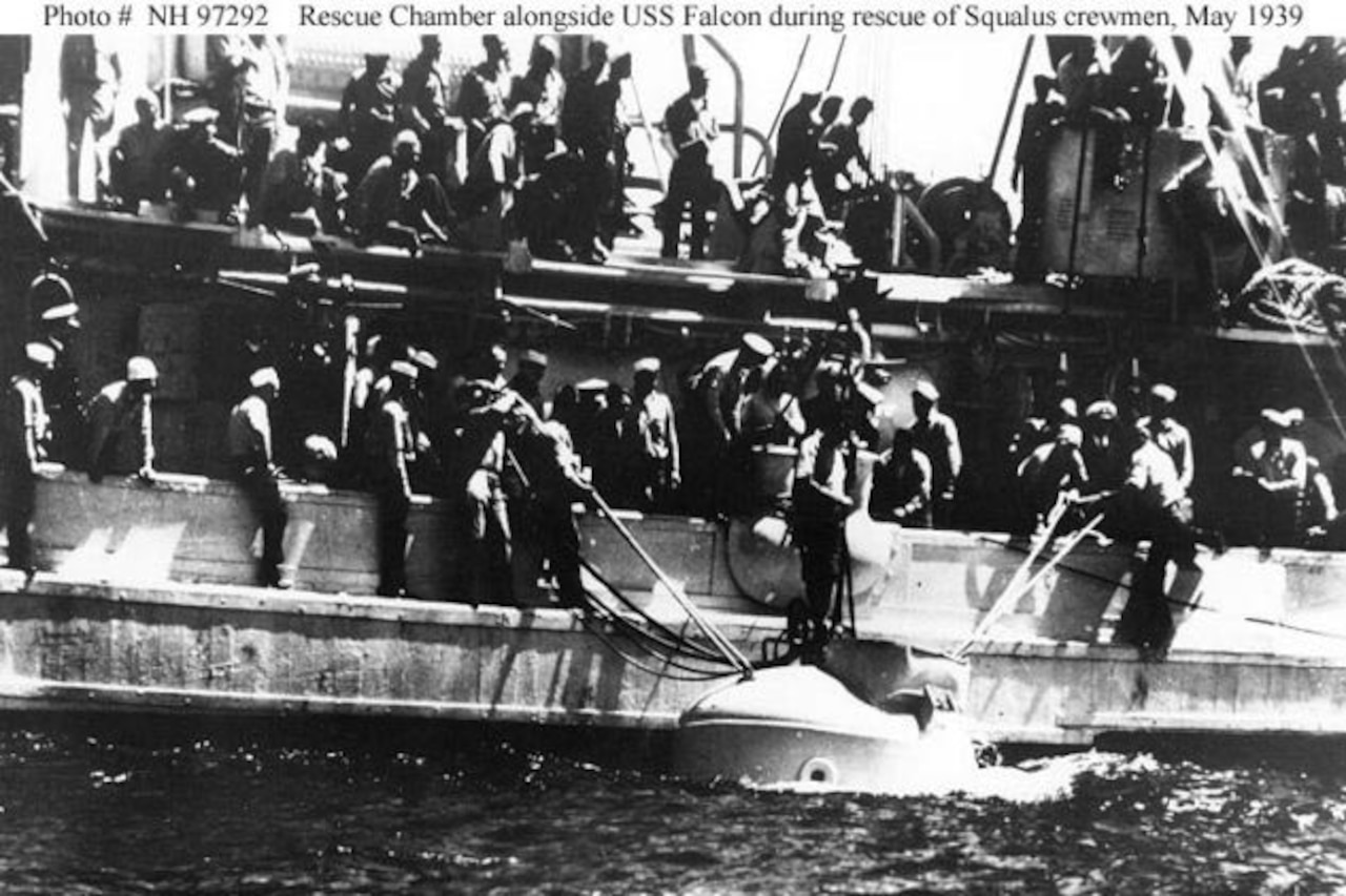 A boat beside a larger vessel surrounded by men pulls in the top of a rescue chamber.
