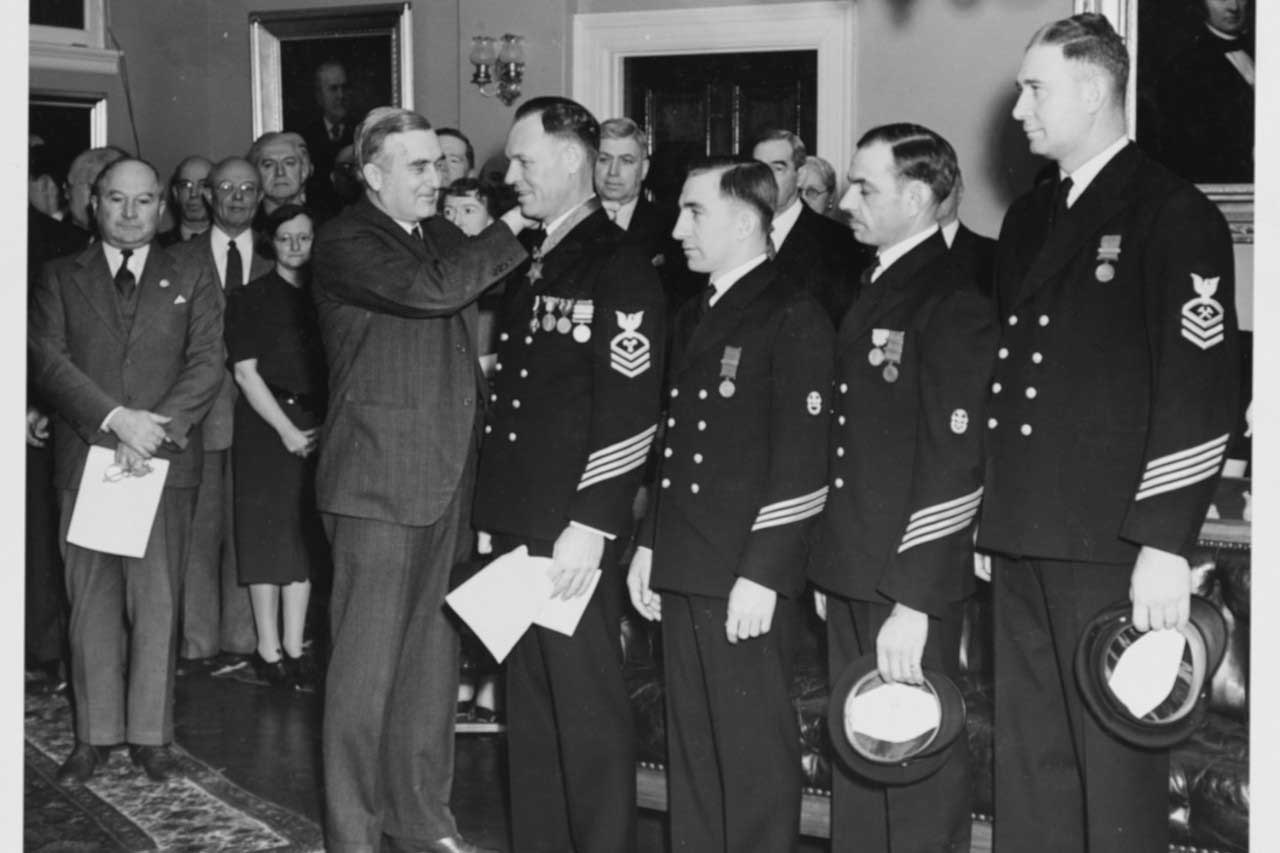 Four sailors in dress blues stand in line to receive the Medal of Honor from a man in a suit as others look on.