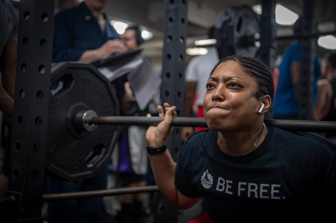 A woman wearing an earbud breathes while holding a barbell behind her shoulders.