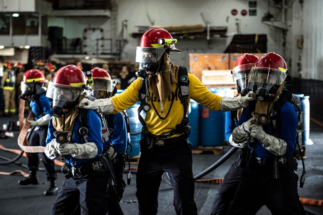 A group of sailors stand in a v formation wearing masks and holding two fire hoses.
