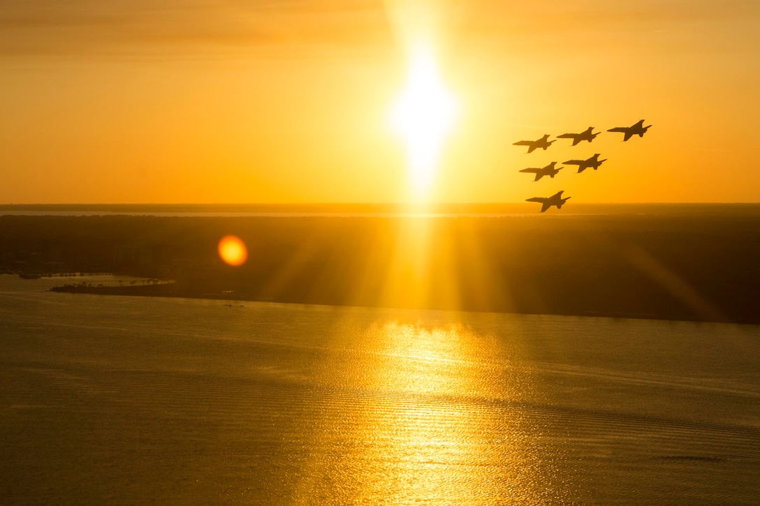 A formation of military fighter jets flies over water with a yellow sky in the background.